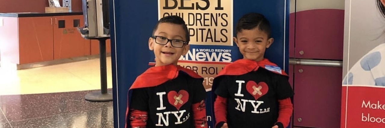 A set of twin boys wearing super hero capes and smiling for the camera.