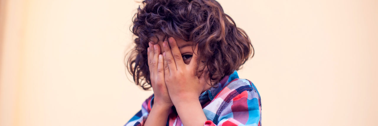 Un niño latino cubre su cara con sus manos. Tiene pelo risado y una camisa rojo y azul.