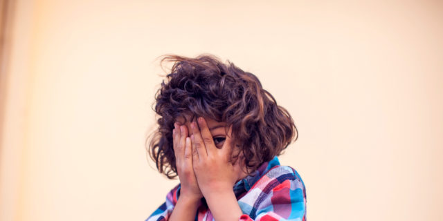 Un niño latino cubre su cara con sus manos. Tiene pelo risado y una camisa rojo y azul.