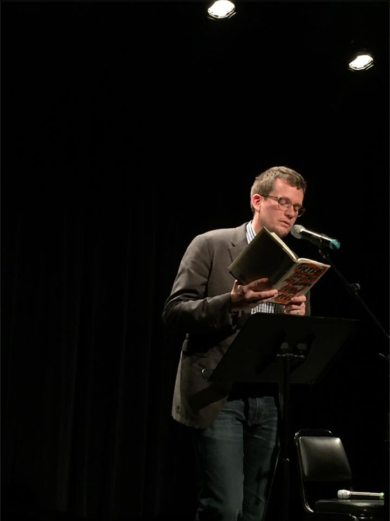 John Green, author, stands and speaks into a microphone, reading from his book. He has brown hair and is wearing a brown coat, white plaid shirt, and blue jeans. He is wearing glasses.