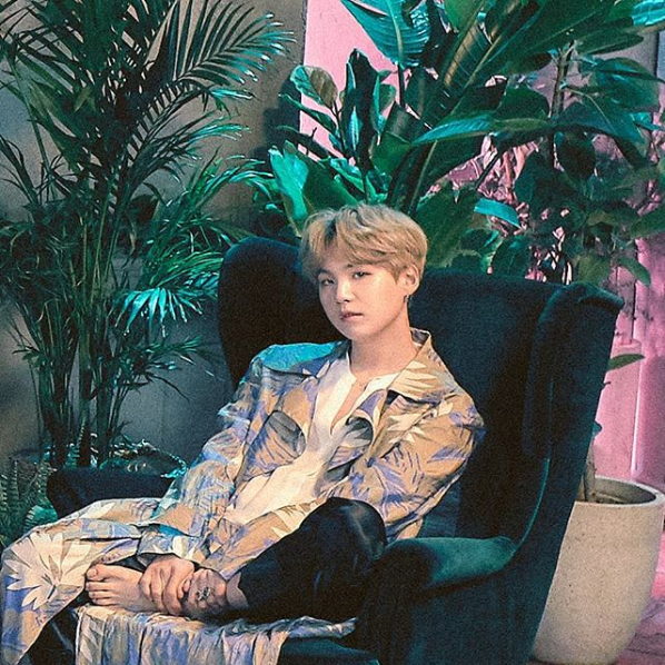 Suga, Korean rapper, sits in a lounge chair. He is wearing a kimono decorated with blue and white palm fronds. He has pink hair.