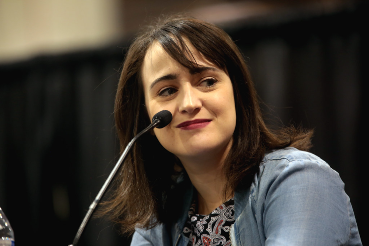 Mara Wilson, writer and performer. She is smiling and leaning into a microphone to answer someone's question. She is wearing a blue jacket. She has long brown hair.