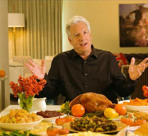 Marc Summers, television host, sits before a large feast. He has white hair. He is wearing a black dress shirt.