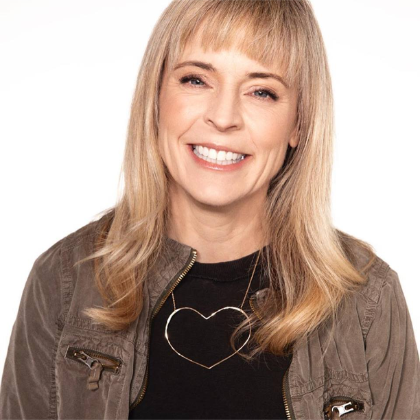 Maria Bamford, comedian. She has long blond hair with bangs. She has a necklace with a heart motif. She is wearing a black shirt and brown coat. She is smiling.