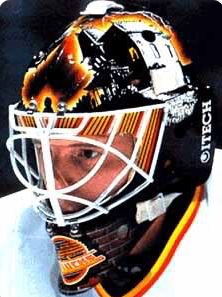 Corey Hirsch, retired NHL player, wearing his helmet. His helmet is black, orange, and yellow. His jersey is white with orange and yellow trim.