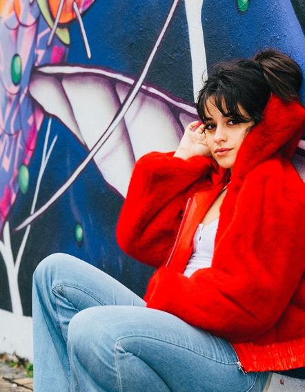 Camila Cabello, kubansk-amerikansk sångerska, poserar framför en väggmålning. Hon bär en röd, fluffig jacka, vit topp och blå jeans. Hon har långt, mörkbrunt hår med pannkaka som hon dragit upp i en hästsvans.