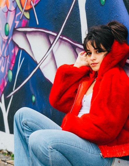 Camila Cabello, Cuban-American singer, poses in front of a mural. She is wearing a red, fluffy jacket, white top, and blue jeans. She has long, dark brown hair with bangs, pulled into a ponytail.