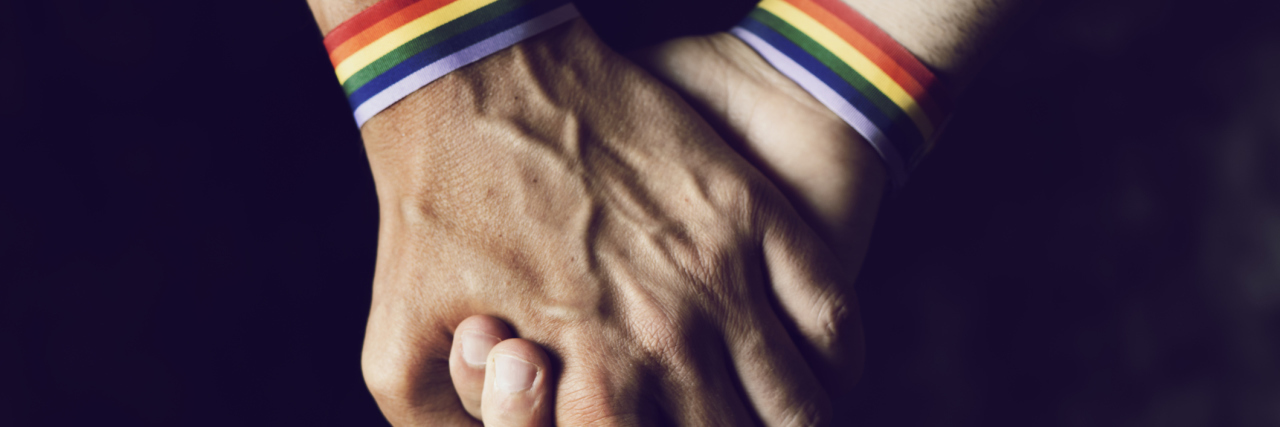 Closeup of two caucasian men holding hands with a rainbow-patterned wristban on their wrists