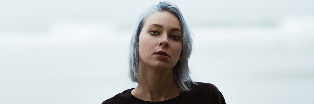 woman with short blue hair and a black sweater standing outside on a cloudy day in front of the ocean. she's looking into the camera with a serious expression.