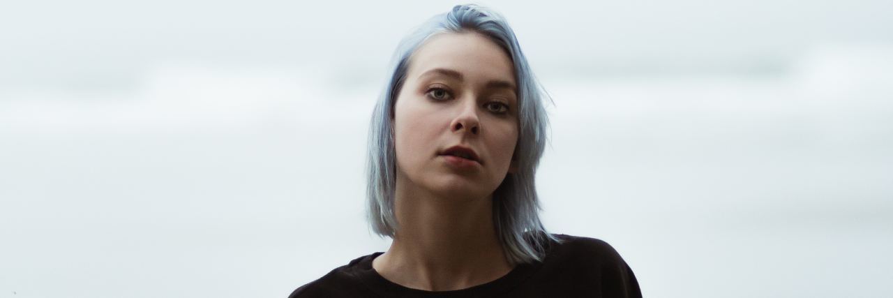 Why You Shouldn't Try to 'Fix' Someone's ME/CFS woman with short blue hair and a black sweater standing outside on a cloudy day in front of the ocean. she's looking into the camera with a serious expression.