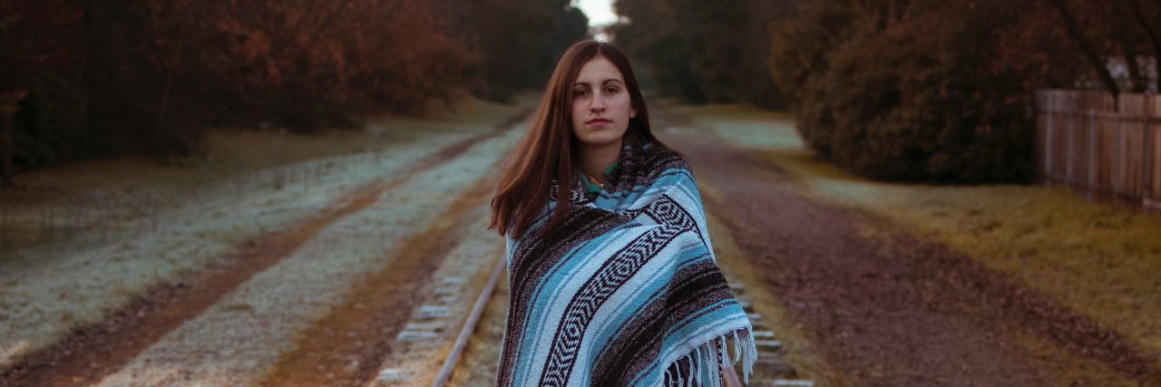 photo of young woman wrapped in blanket standing in middle of train tracks and looking at camera