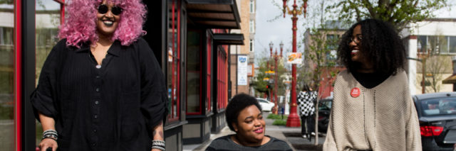 Three Black and disabled people smiling at each other while strolling down a sidewalk side by side. On the left, a non-binary person walks with a cane in one hand and a tangle stim toy in the other. In the middle, a woman rolls along in her power wheelchair. On the right, a woman walks and gestures.