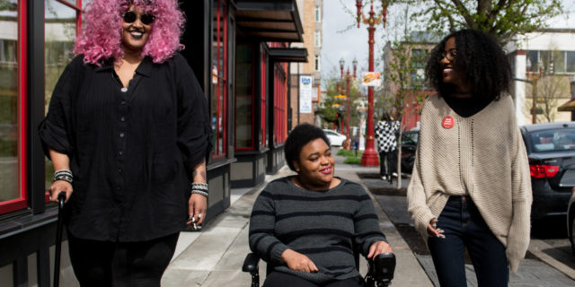 Three Black and disabled people smiling at each other while strolling down a sidewalk side by side. On the left, a non-binary person walks with a cane in one hand and a tangle stim toy in the other. In the middle, a woman rolls along in her power wheelchair. On the right, a woman walks and gestures.