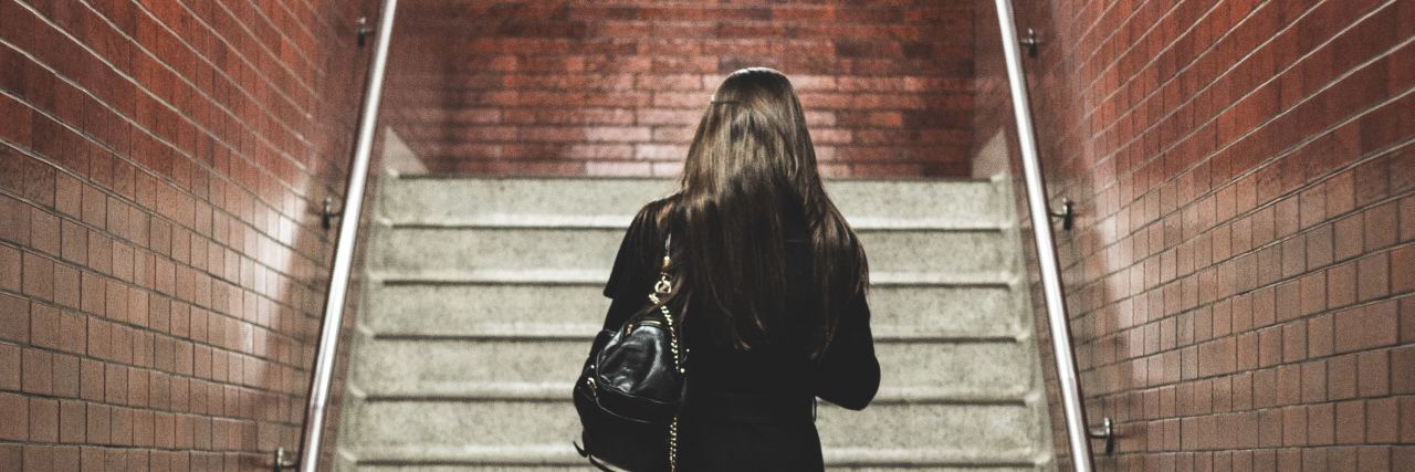 I Quit My Job for My Mental Health — and I Feel Good About It back of a woman in a black dress with a purse over her shoulder about to walk up a flight of stairs