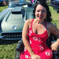 Mollie wearing a red sundress, sitting in her power wheelchair outdoors at a car show.