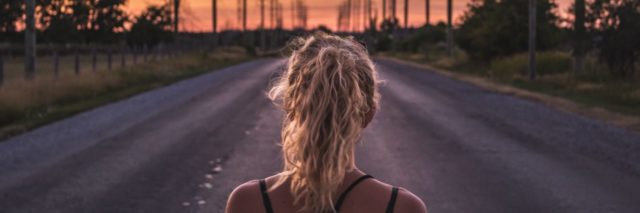 woman standing in middle of road looking toward horizon at sunset