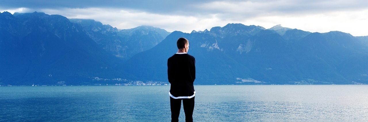 photo of man standing by lake shore alone with mountains in distance