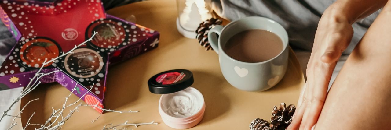 photo of assortment of self-care goods on wooden tray on bed with a woman's legs in shot, including tea, treats and lotions