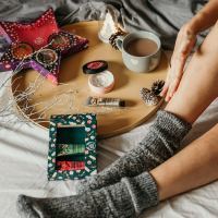 photo of assortment of self-care goods on wooden tray on bed with a woman's legs in shot, including tea, treats and lotions