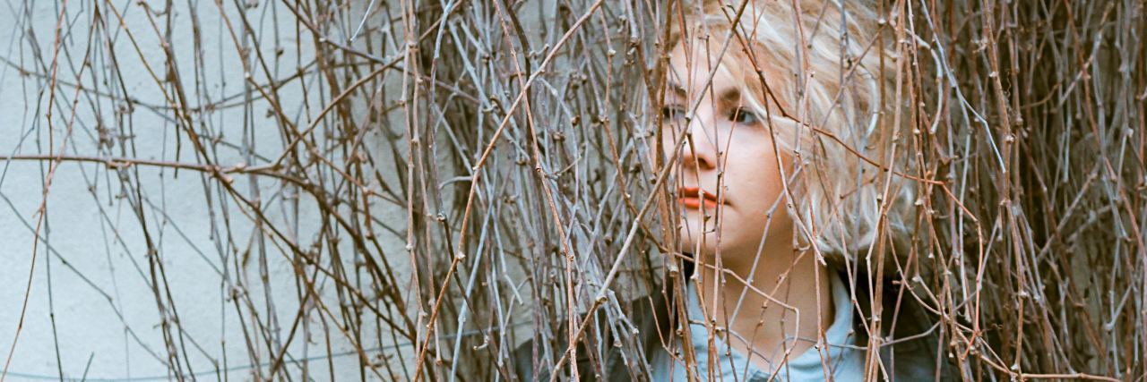 What It Means to Be 'Addicted' to Self-Harm woman standing in layers of brig and branches looking off-camera