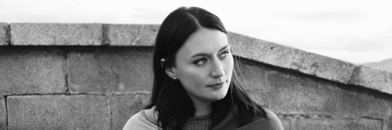 Why People With Chronic Illness Often Experience Imposter Syndrome Jennifer leaning back against a wall, wrapped in a blanket.