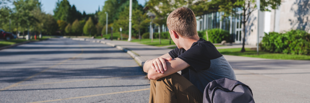 The School Challenges My Son Faces Due to ADHD, Anxiety and Depression Boy sitting on the curb in front of his school looking away from camera