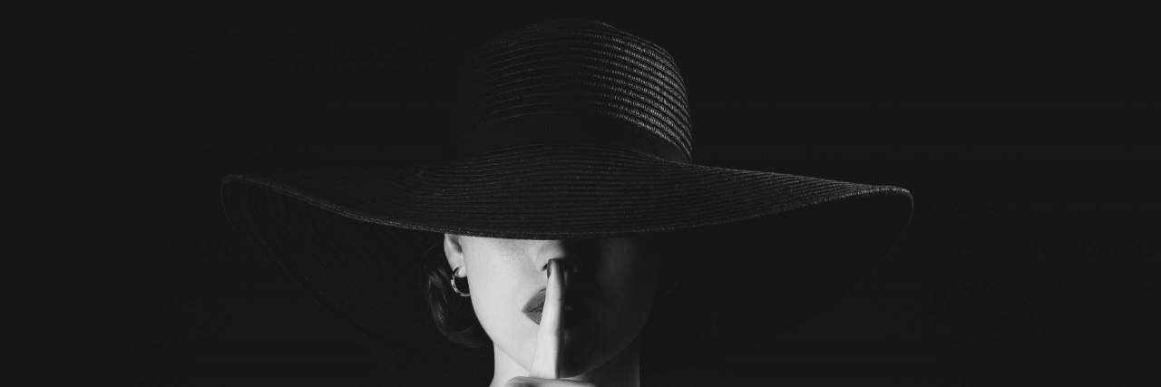 What to Tell Family and Friends About Living With Chronic Illness woman with a finger in front of her lips in a large hat covering her eyesin black and white