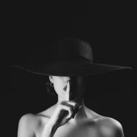 woman with a finger in front of her lips in a large hat covering her eyesin black and white
