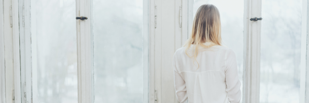 What It's Like Living With Complex PTSD From Childhood Trauma A rear view of a blonde woman in a white shirt standing by a window.