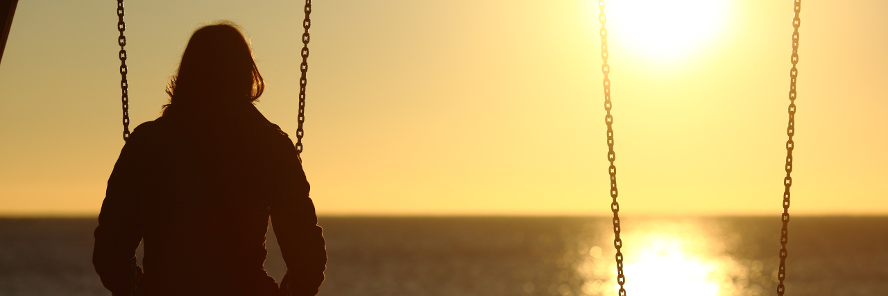 woman on a swing with her back toward the camera looking into a sunset over the beach with another empty swing next to her