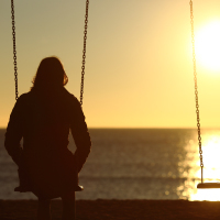 woman on a swing with her back toward the camera looking into a sunset over the beach with another empty swing next to her