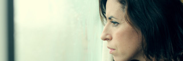 A woman stands and intensely stares out of a window.