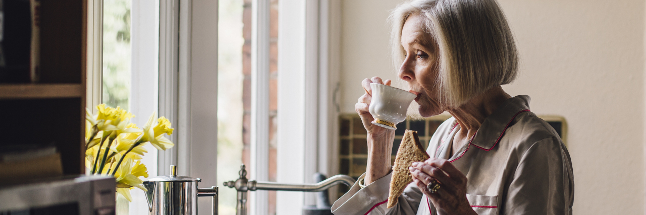 photo of older woman eating toast and drinking coffee while looking out window