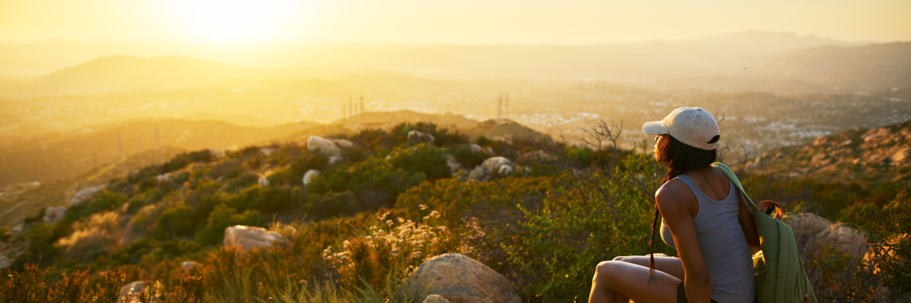 Rear view of a woman with a white hat and blue tank top sitting on a rock on top of a hill overlooking the sunset and greenery