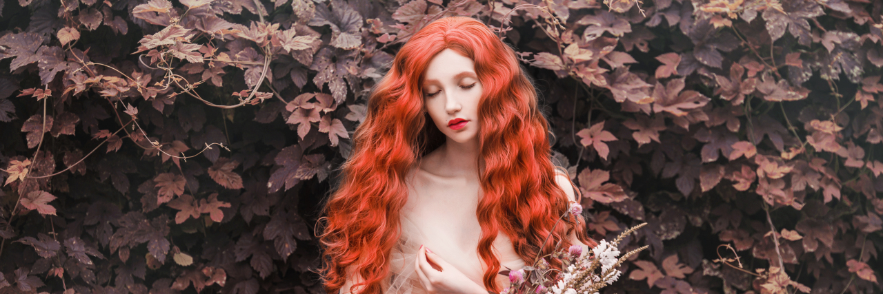 Too Many Women With Chronic Illnesses Are Disbelieved and Dismissed Woman with curly red hair with a tulle dress in a garden, holding flowers.