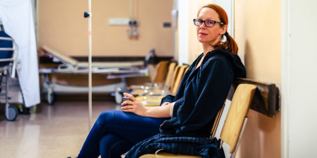 Patient sitting in hospital ward hallway waiting room with iv. Woman with intravenous therapy in her hand is waiting in the clinic corridor with blurred medical personnel in background.