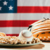 Baseball on plate with peanuts, leather glove and hot dogs on table with us flag behind.