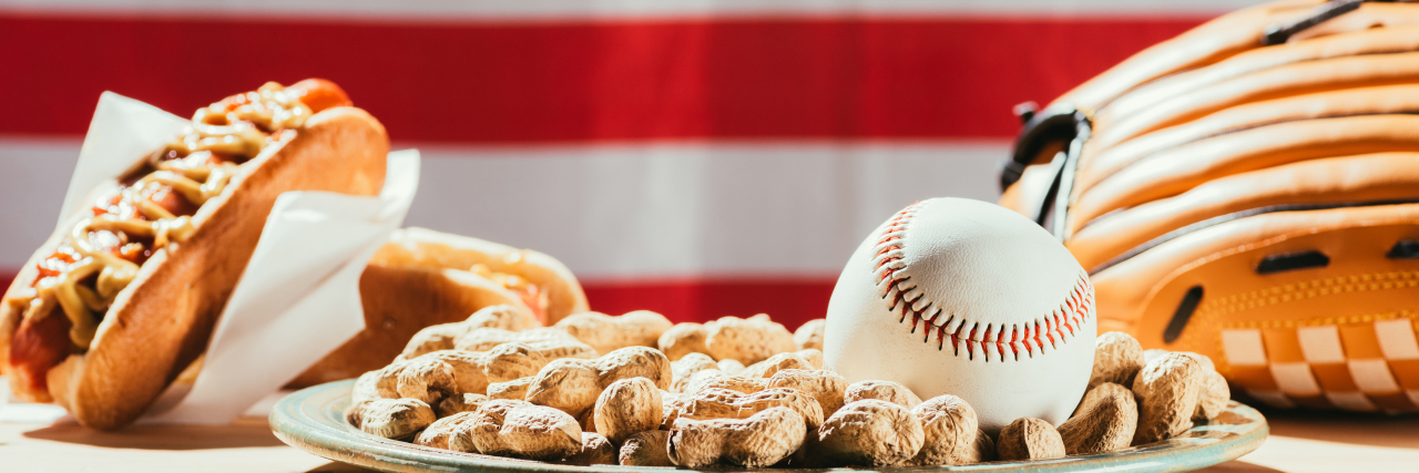 What a Ball Game Taught Me About Inclusion and the Meaning of 'Normal' Baseball on plate with peanuts, leather glove and hot dogs on table with us flag behind.
