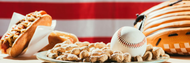 Baseball on plate with peanuts, leather glove and hot dogs on table with us flag behind.