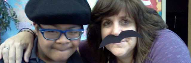 Selfie of child with a disability and teacher. Teacher is wearing a fake mustache.