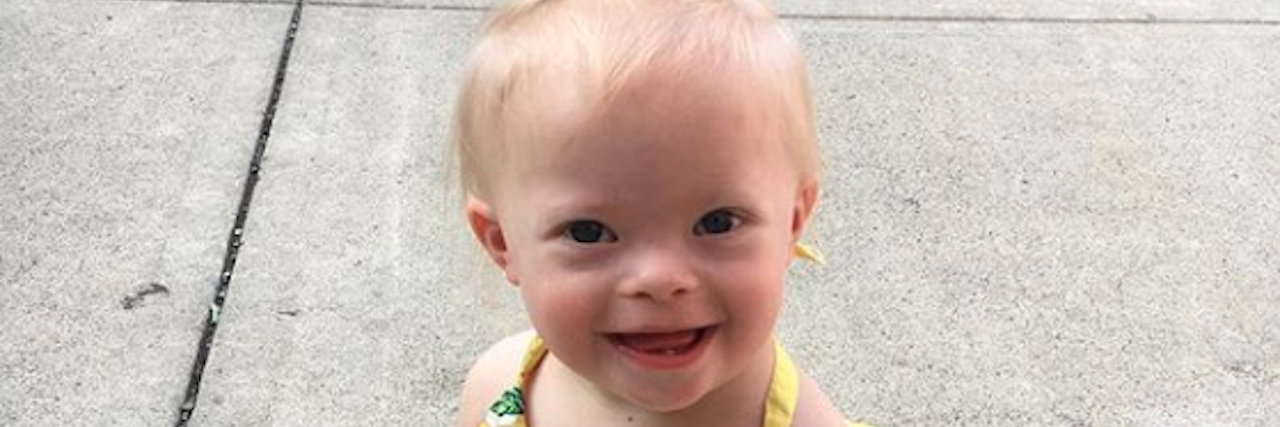 What I Wish Doctors Said When Delivering a Down Syndrome Diagnosis Little girl with Down syndrome sitting on floor smiling at camera