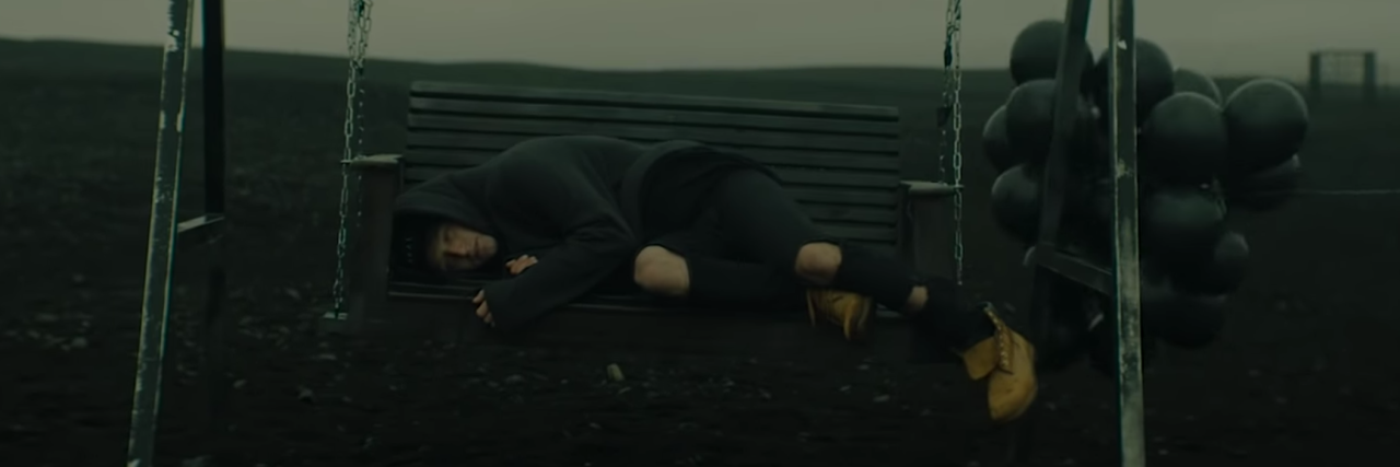 screenshot from NF song The Search, with rapper lying on swing on desolate landscape with black balloons blowing around him