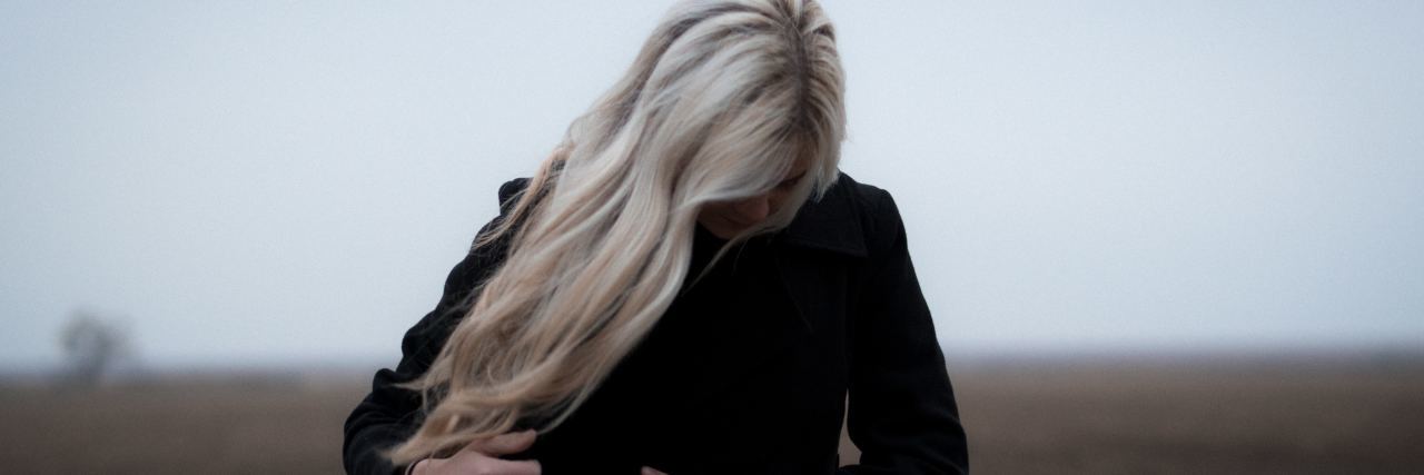 A woman with blonde hair and a white coat looking down. She stands outside in front of a bleak landscape.