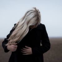 A woman with blonde hair and a white coat looking down. She stands outside in front of a bleak landscape.