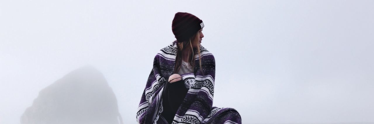 Girl on top of a car wrapped in a blanket looking ro the right. White background, red beanie, long hair