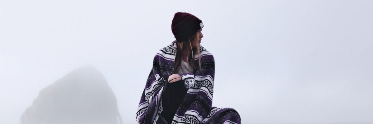 How I Manage My Mental Health With Crohn's Disease Girl on top of a car wrapped in a blanket looking ro the right. White background, red beanie, long hair