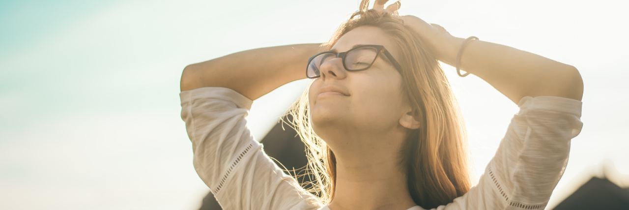 Realizing My Chronic Migraine Is Not My Fault photo of blonde woman standing in sunshine with eyes closed and hands on her head
