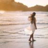 photo of woman in white dress walking alone beach in golden light