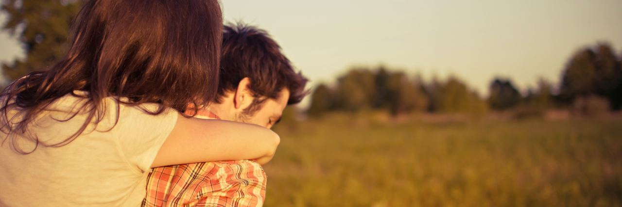 photo of woman with arms around man from behind while he carries her over a sunny field