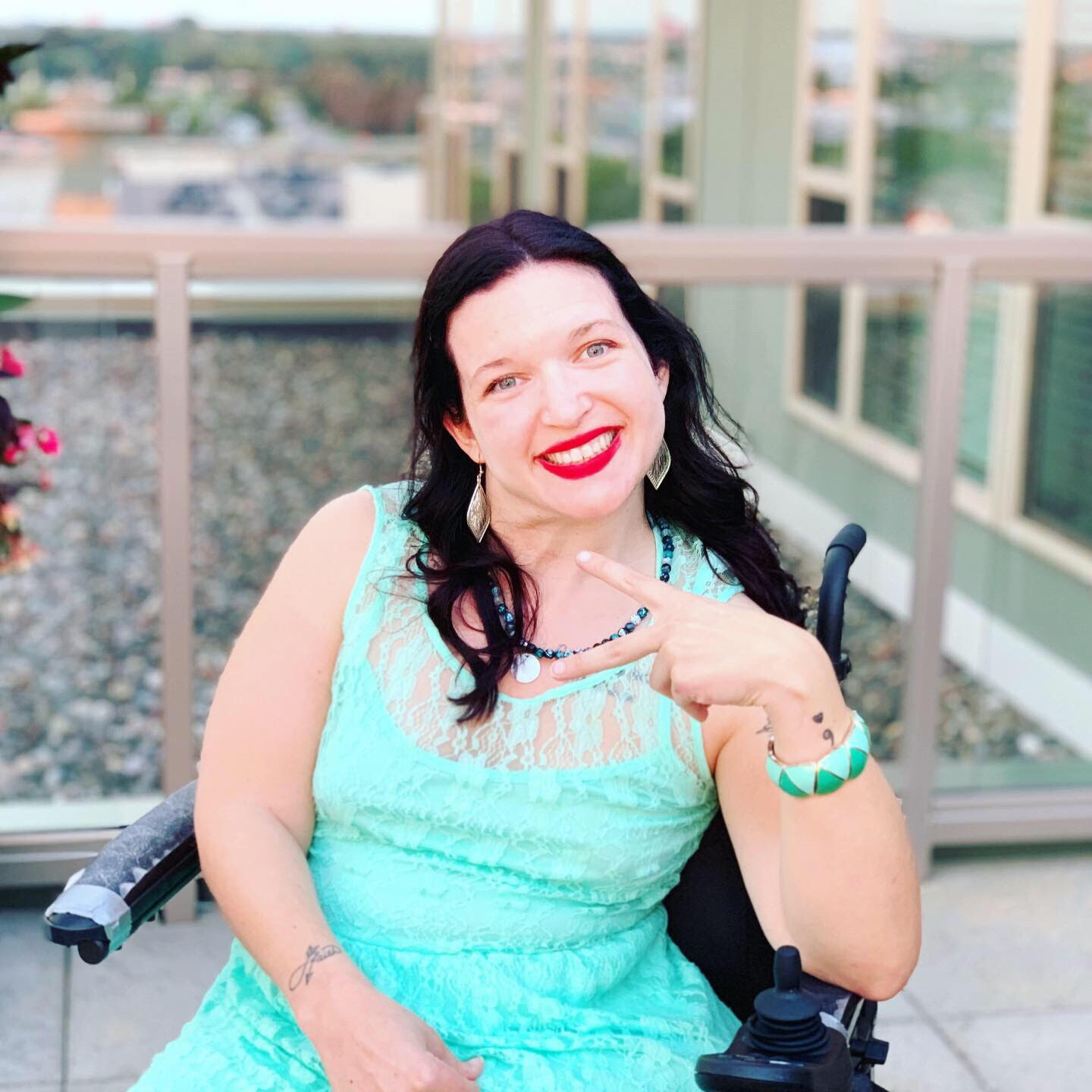 Mollie making the peace sign. She is sitting in her power wheelchair and wearing a bright turquoise dress.
